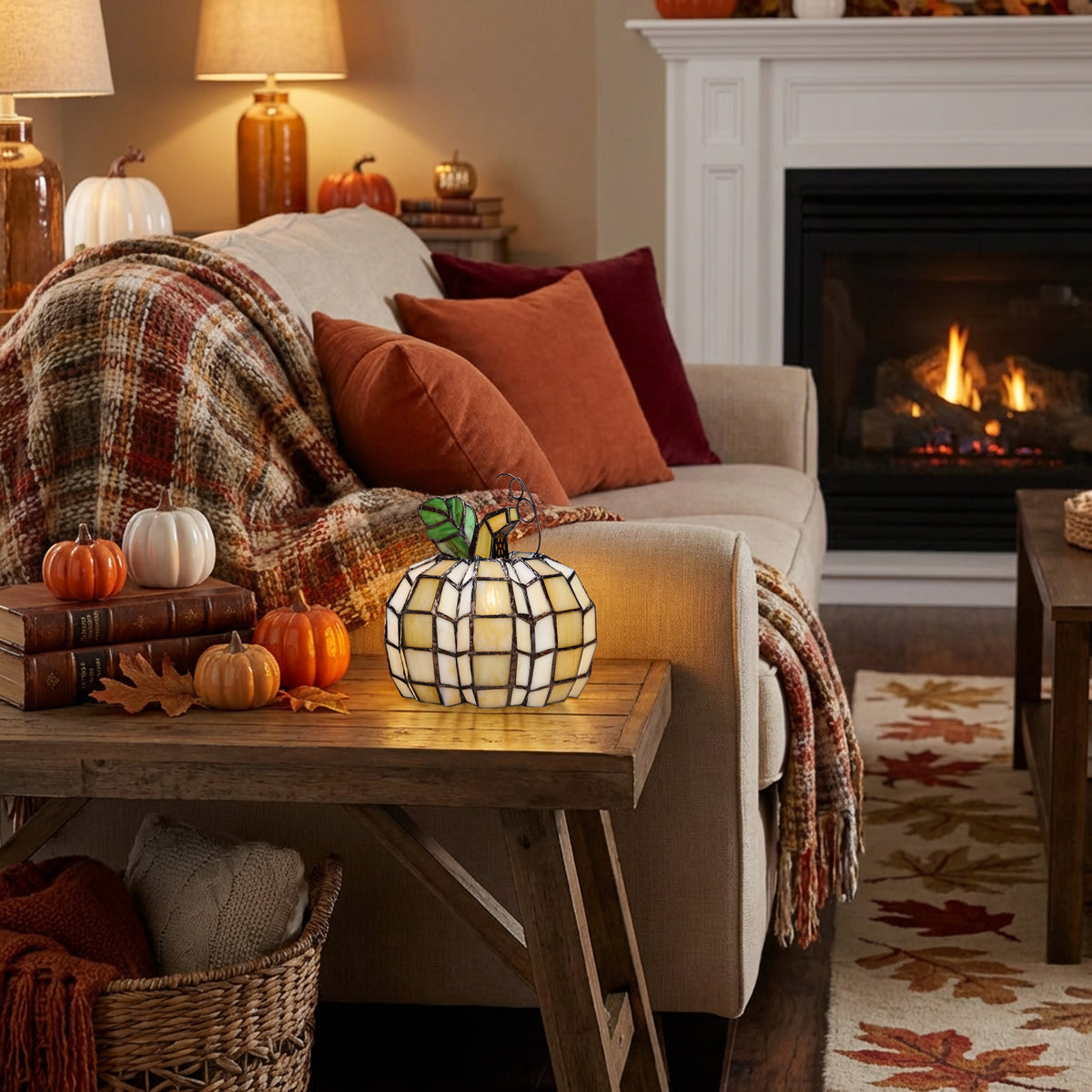 9"H Patch the Pumpkin Stained Glass Accent Lamp sits on a wooden coffee table in a cozy living room. A beige sofa with rust and burgundy pillows, a plaid throw blanket, and small pumpkins are arranged nearby, with a fireplace and warm lamps in the background.