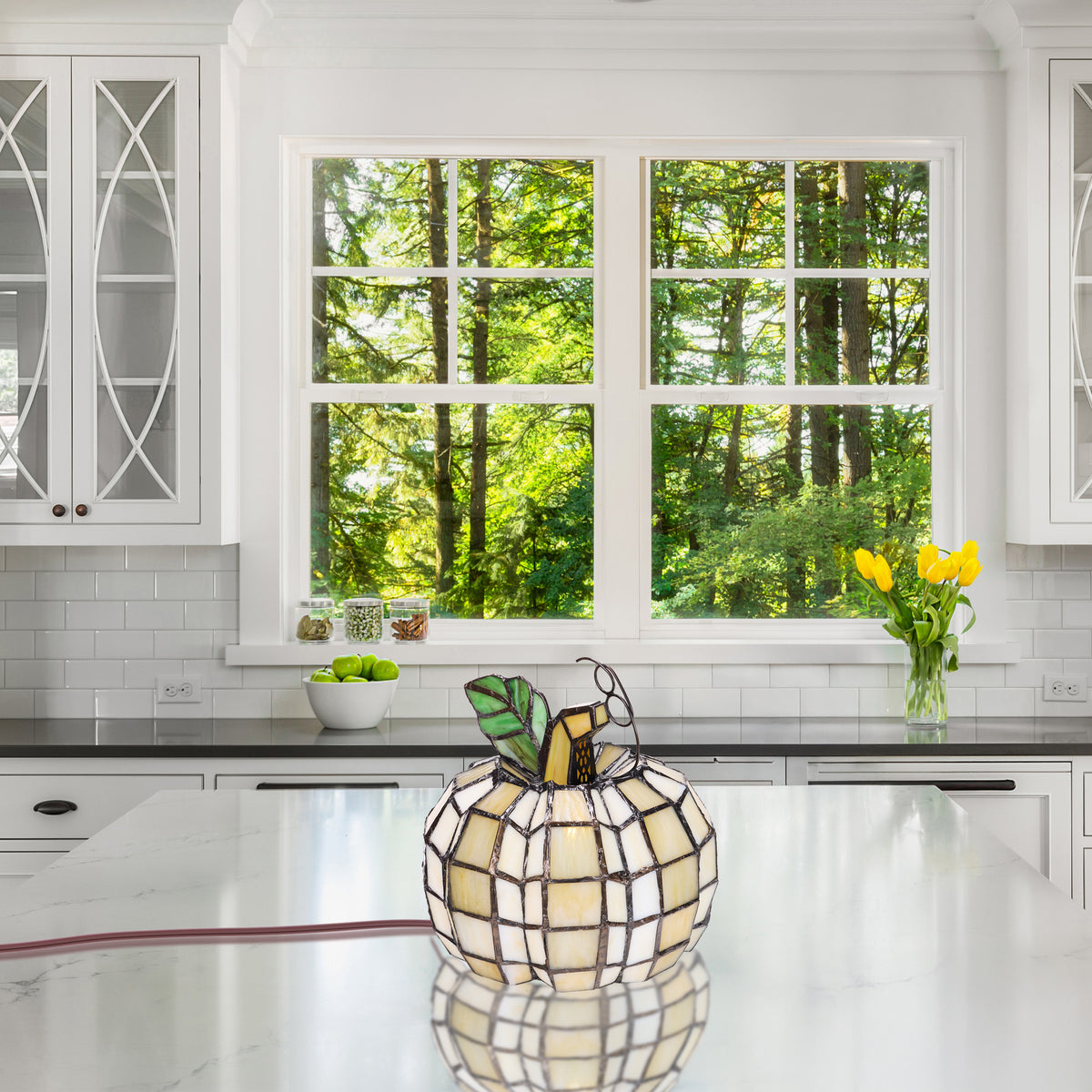 9"H Patch the Pumpkin Stained Glass Accent Lamp sits on a white marble kitchen island, with a bright modern kitchen and a window showing green trees in the background.